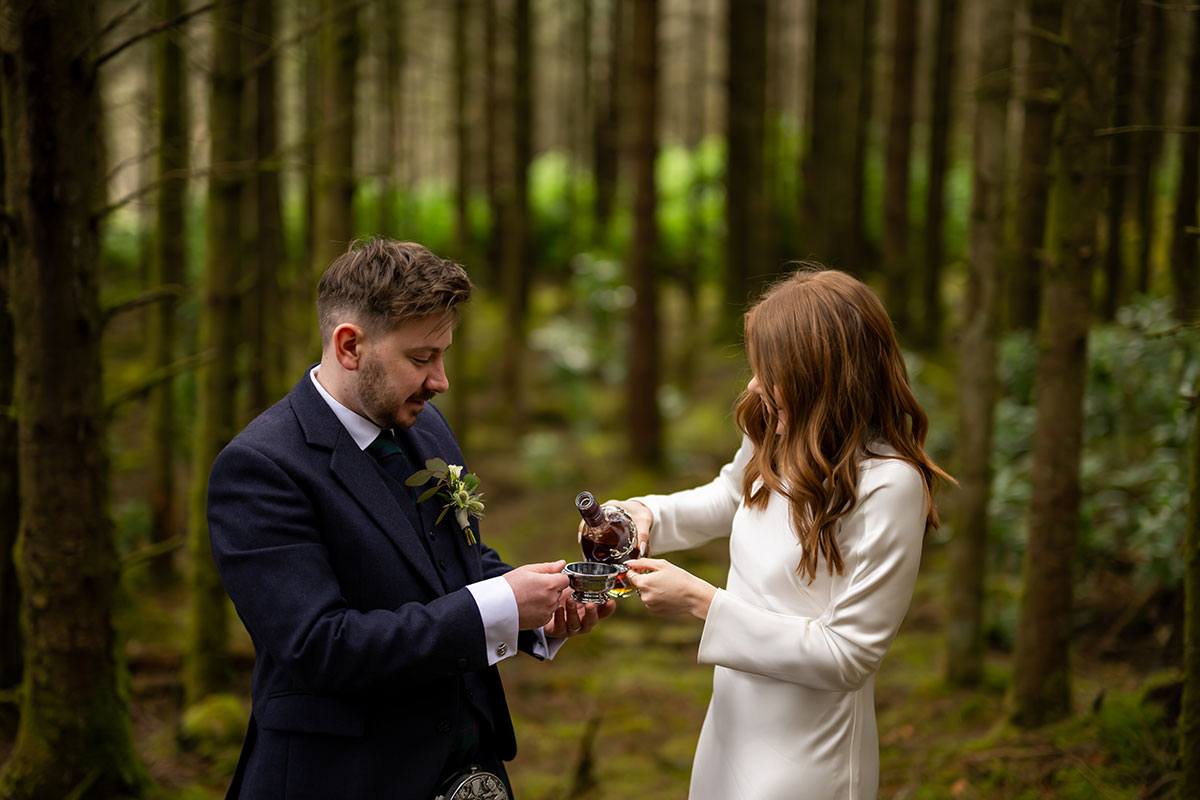 Bride pours whisky into a quaich during an outdoor Scottish wedding ceremony in a woodland setting in Glencoe
