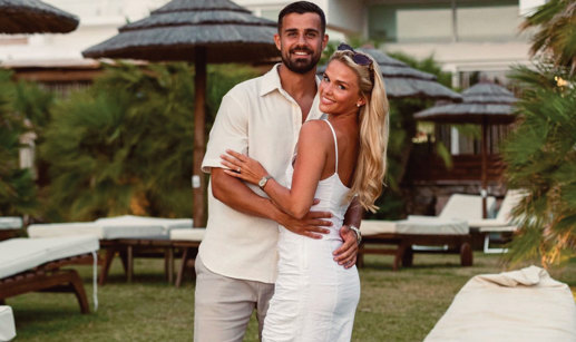 a smiling man and woman posing for a photograph in front of sunbeds and sun parasols with greenery in the background