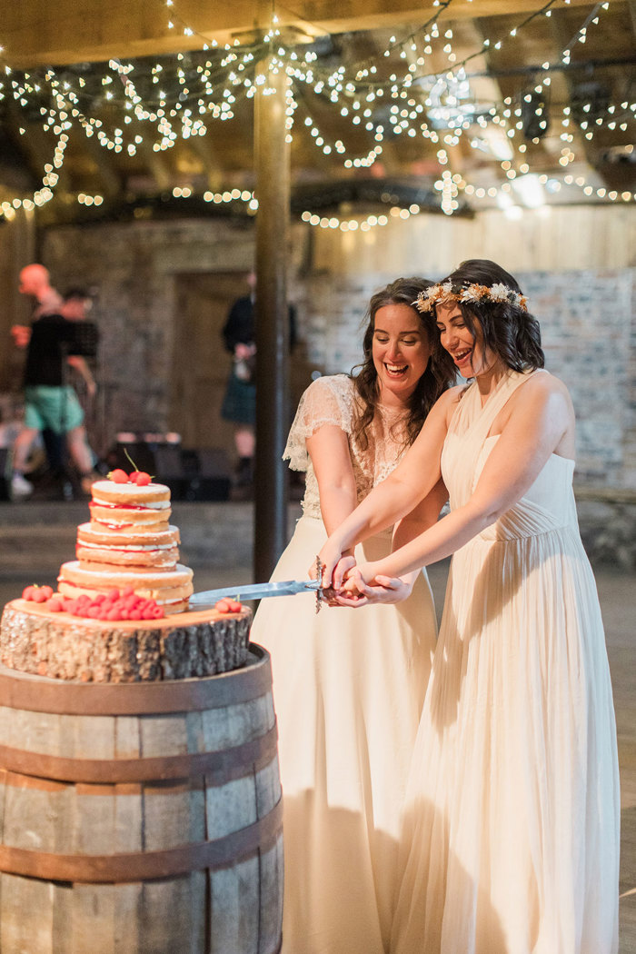 two joyous brides cutting a stack of cakes with a sword. The cake is sitting on a large wooden barrel in a room with exposed brick walls and fairy lights overhead.