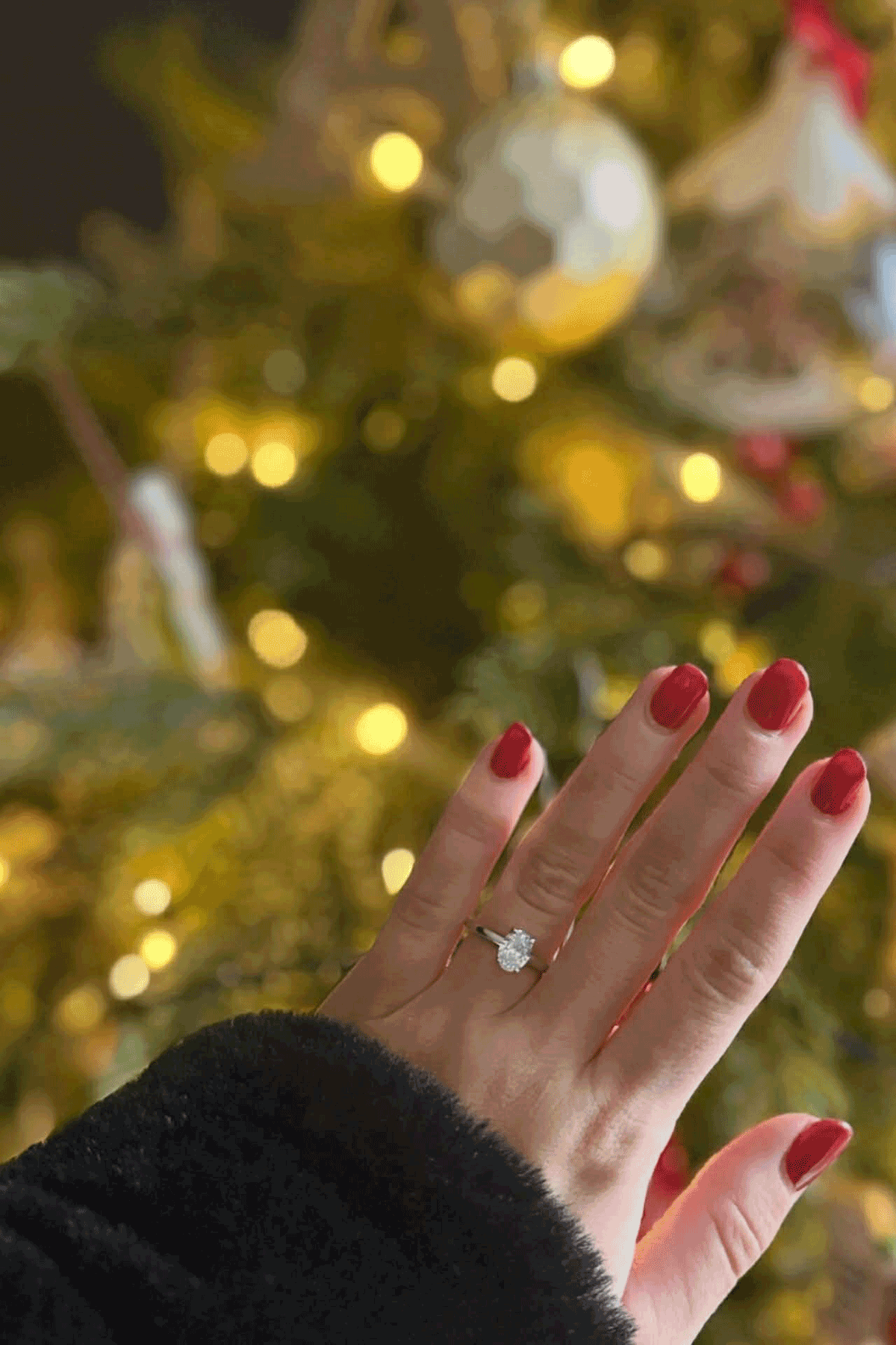 Close-up of diamond engagement ring on hand with red nails against festive Christmas tree lights