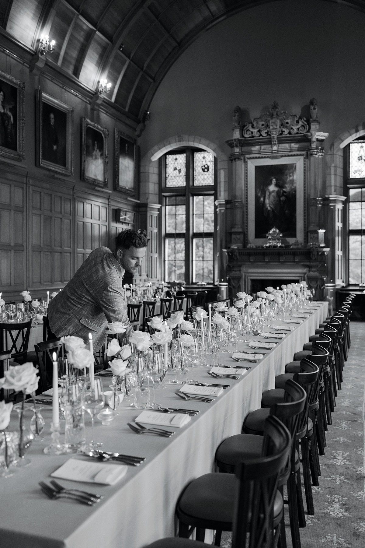 Wedding planner adjusting long dining tables styled with white roses, candles and place settings inside an elegant historic dining hall