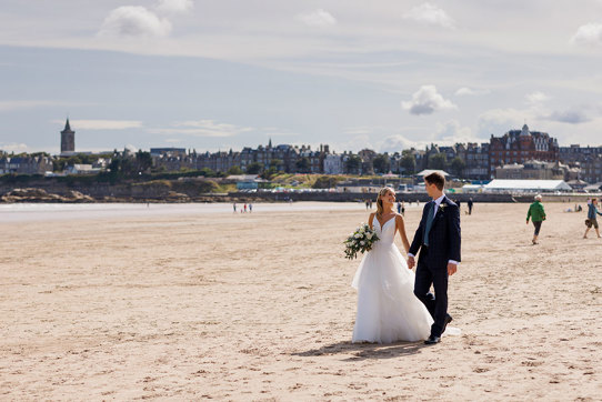 A newlywed couple holding hands and walking across a sandy beach, after their Old Course Hotel wedding, with St Andrews and cloudy blue sky in the backdrop.