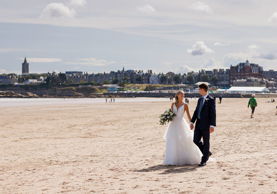 A newlywed couple holding hands and walking across a sandy beach, after their Old Course Hotel wedding, with St Andrews and cloudy blue sky in the backdrop.
