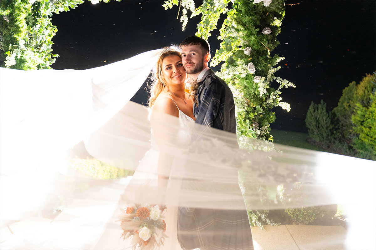 A bride and groom smile at the camera as the bride's veil is swept around them dramatically