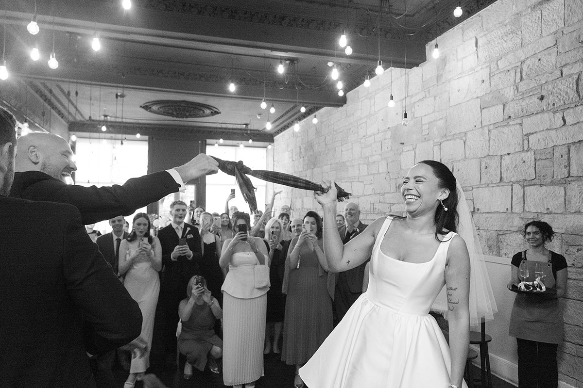 Bride and groom laughing during handfasting ritual with tartan cloth at Glasgow wedding reception with guests watching