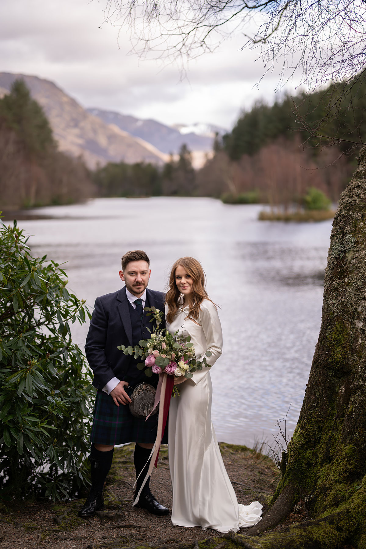 Bride and groom posing beside a loch in Glencoe after their Scottish elopement, with mountain views and a romantic bridal bouquet