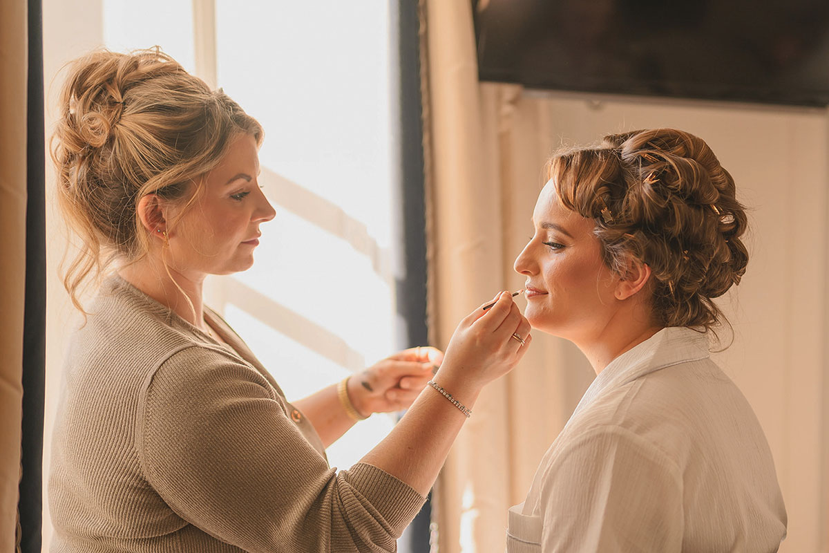 Makeup artist applying lipstick to a bride seated by a window while her hair is styled in an intricate updo