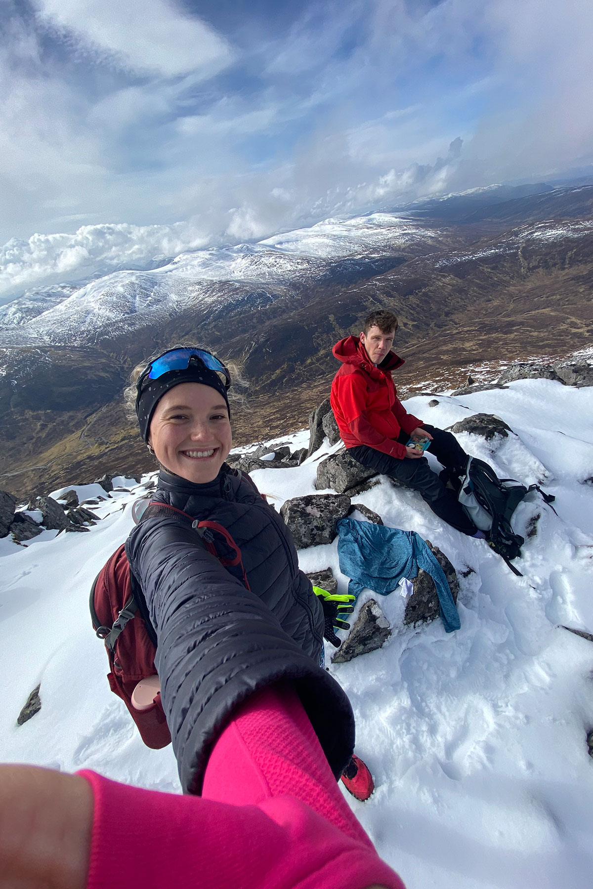 Hikers taking a selfie on a snowy Scottish mountain summit with dramatic Highland landscape and snow-covered peaks behind them