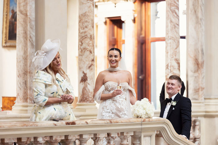 a person wearing a floral dress and a hat making a speech as a bride and groom watch on smiling.