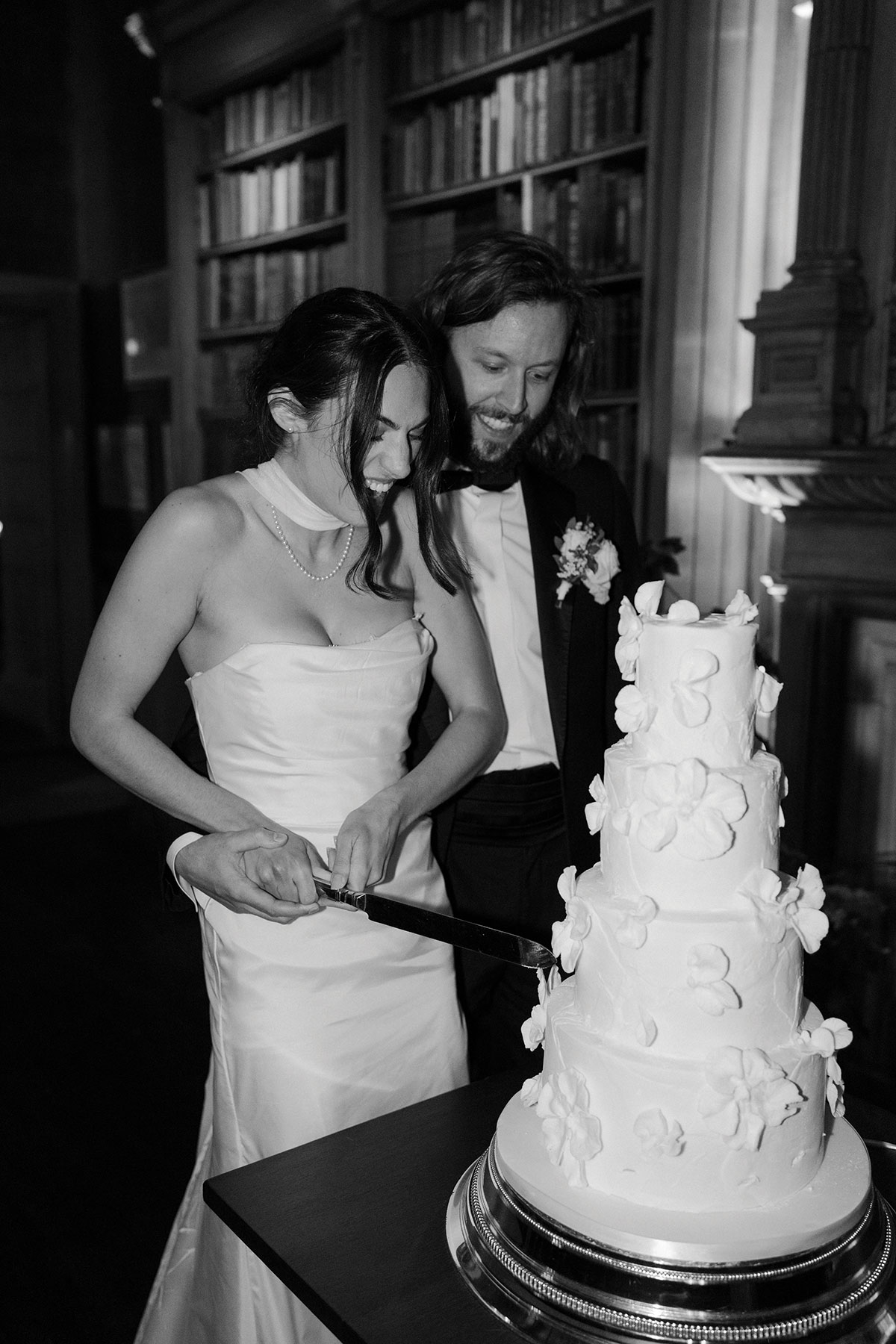 Bride and groom cutting a multi-tier white wedding cake decorated with delicate floral details, photographed in black and white