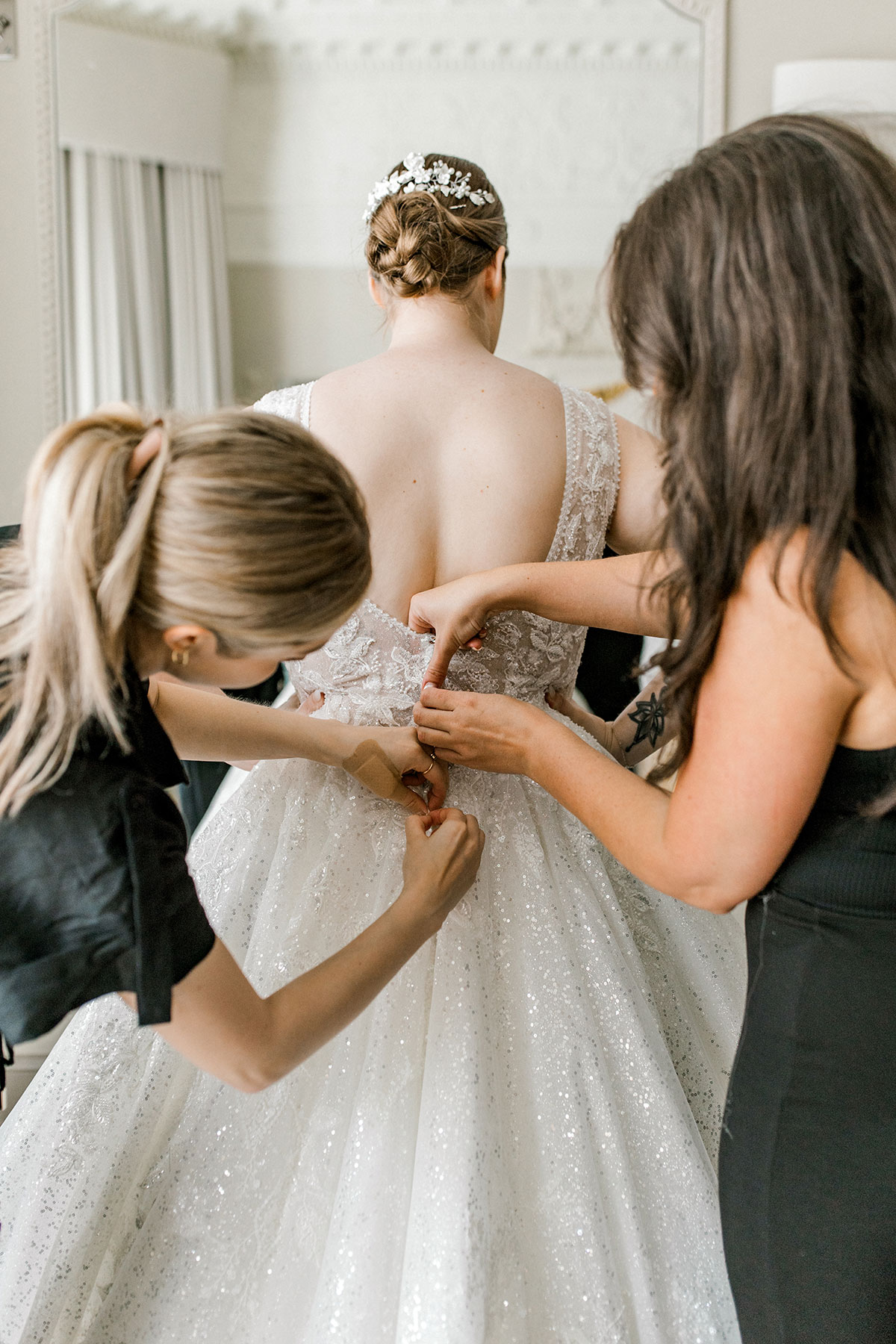 blonde and brunette women help button up the back of bride's wedding dress, one of them stood on either side