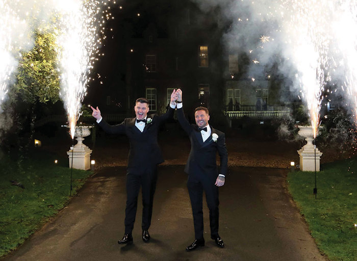 two grooms raise held hands in air outside Carlowrie Castle at night as pyrotechnic flares light up either side of them