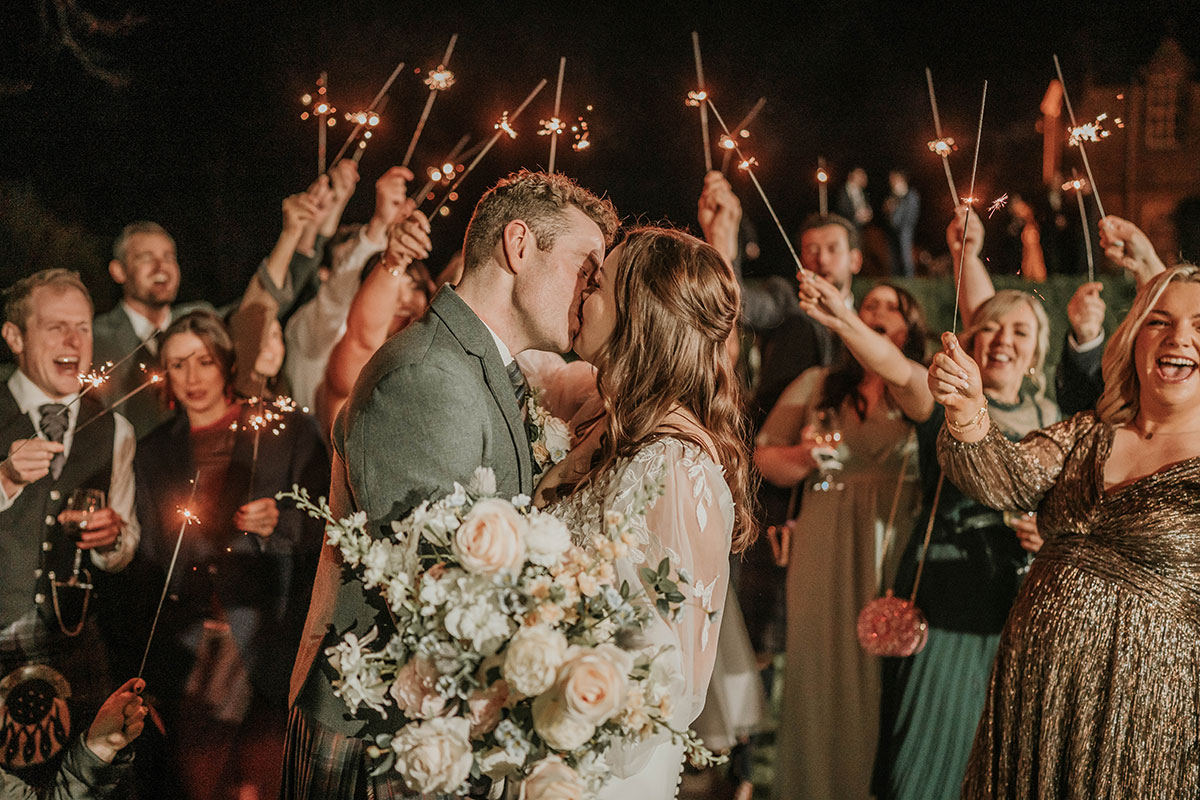 Evening sparkler image with the bride and groom kissing surrounded by cheering guests
