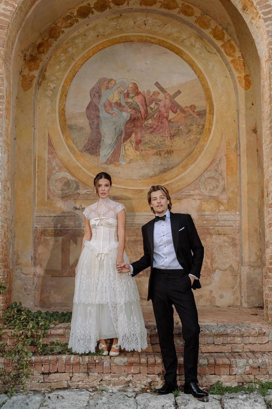 brunette woman in cream corset lace neck and sleeve wedding dress stands on a brick step, one higher than man in tux holding her hand, both in front of religious wall mural