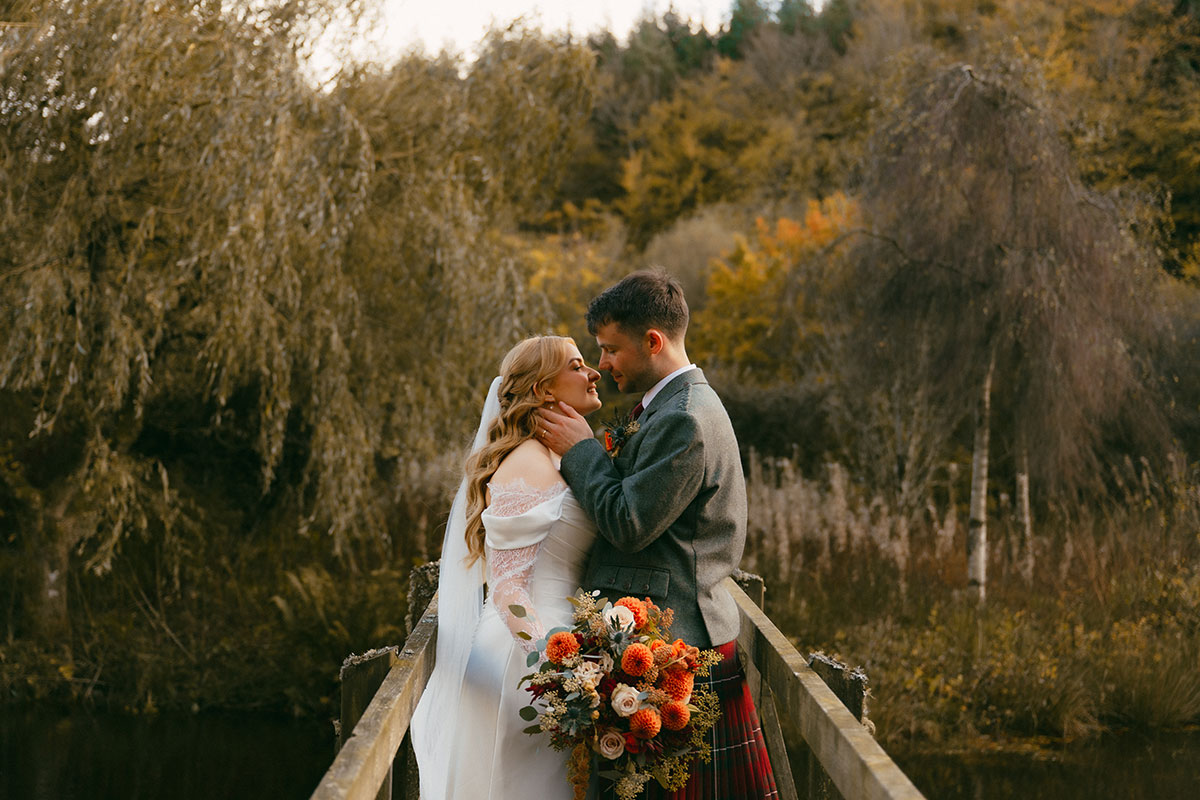 Bride and groom embracing on wooden bridge with autumn foliage and orange bouquet at Scottish woodland wedding