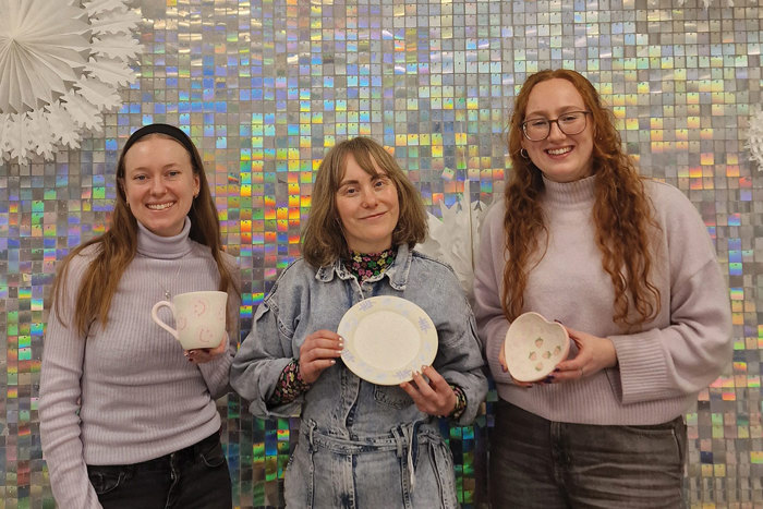 three women stand in front of a shiny disco tiled wall, each smiling and holding up their original painted pottery pieces