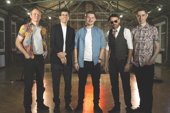 Five men wearing black trousers and colourful tops smiling and standing in a row