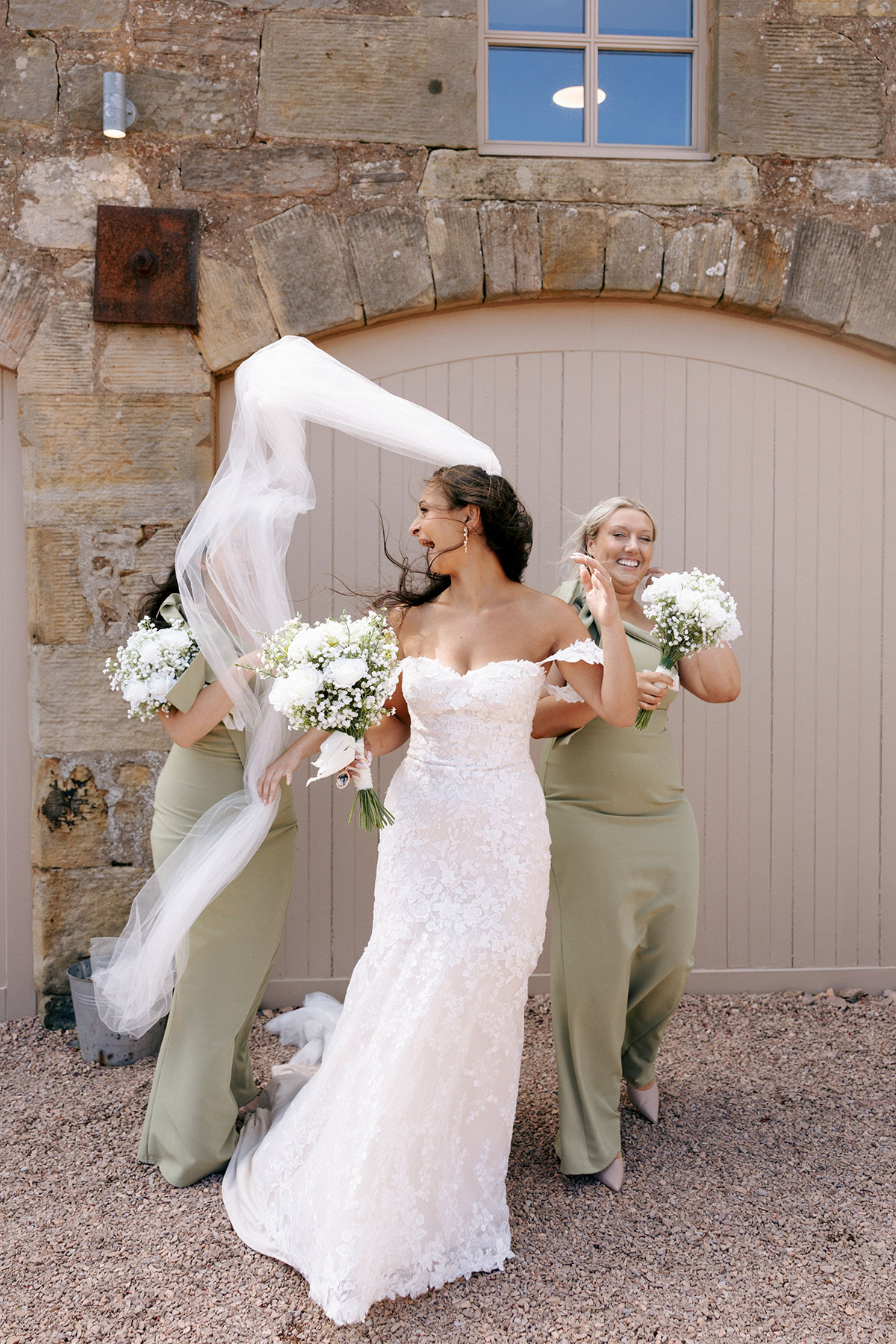 Bride laughs as wind catches her veil outside Falside Mill with bridesmaids in sage green dresses