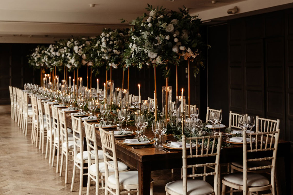 long wooden dining table set up with tall floral centrepieces and formal wine glass place settings and candlesticks