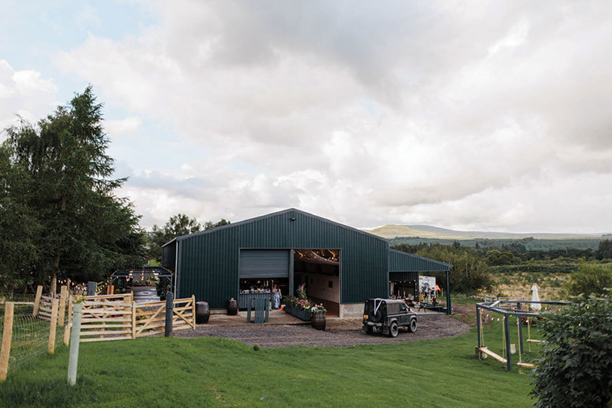 a black painted barn, surrounded by grass and fields beyond, has two open doors and fairylights strung inside 