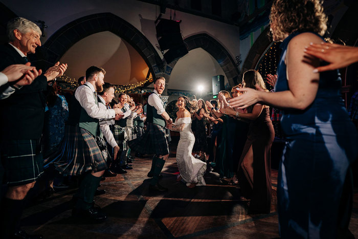 large dimly lit room where two rows of people in occasion wear stand and clap on either side of a bride in a wedding dress and groom in a kilt outfit as they hold hands and skip down the middle