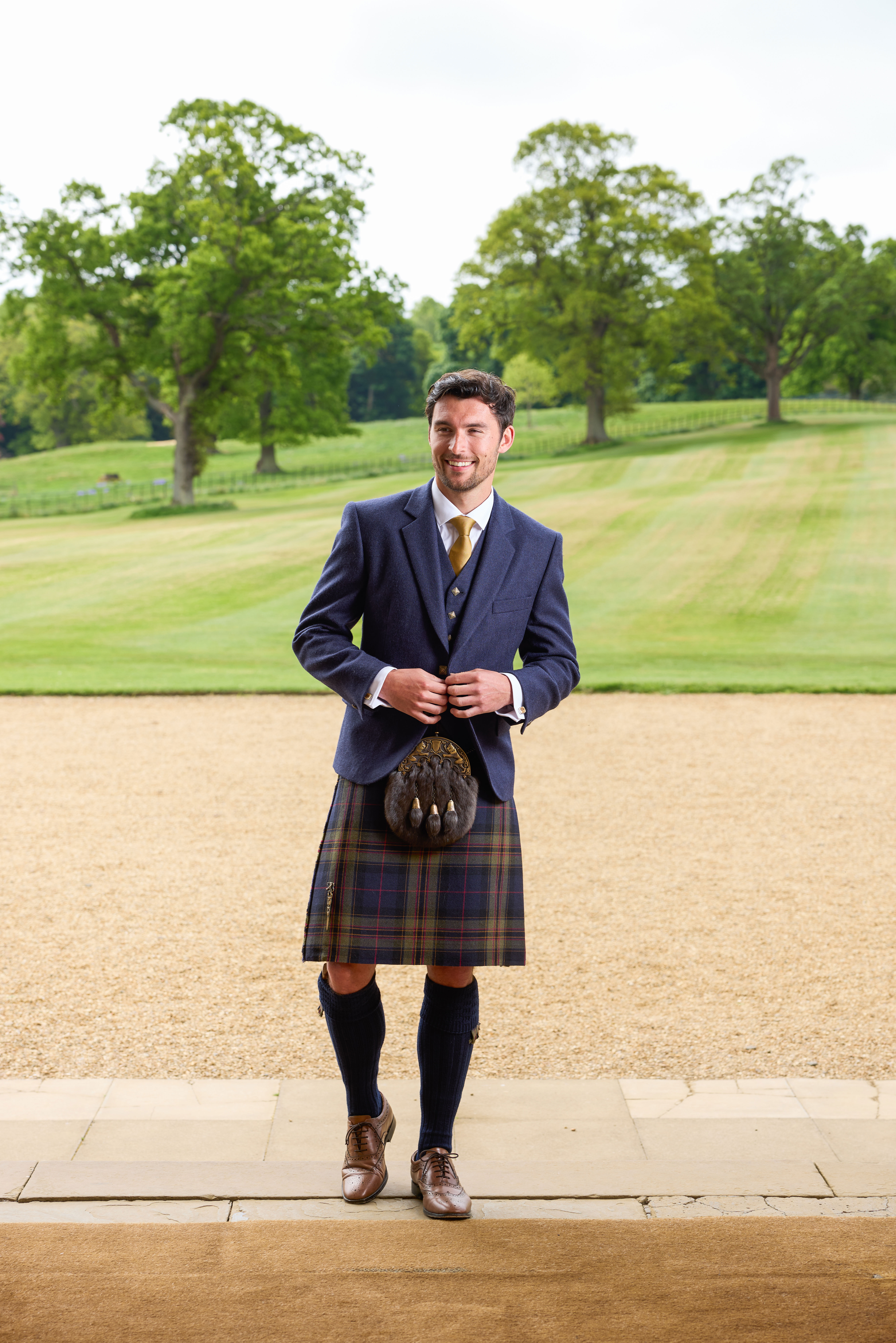 groom smiles while wearing mccalls highlandwear's new noble print tartan kilt with green grass and trees behind him