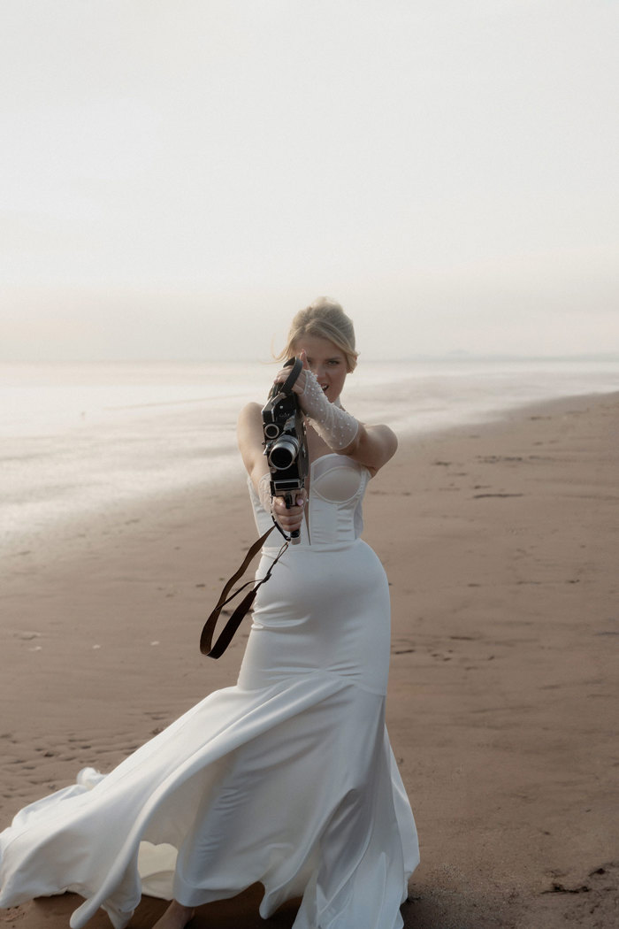 woman in corset wedding dress stands on beach with her hair up, holding an old looking camera out in front of her