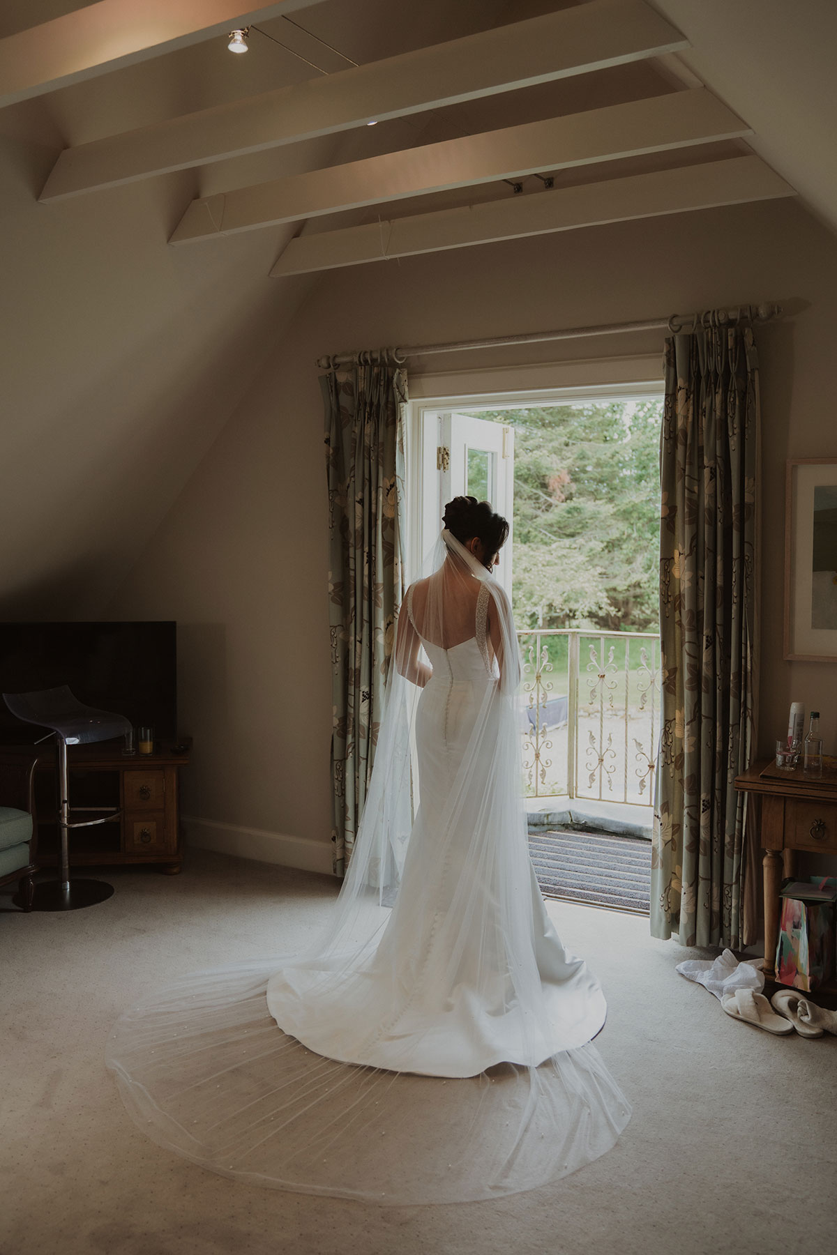 Bride in fitted satin wedding dress and long veil standing at balcony doors during morning preparations at Scottish country house venue