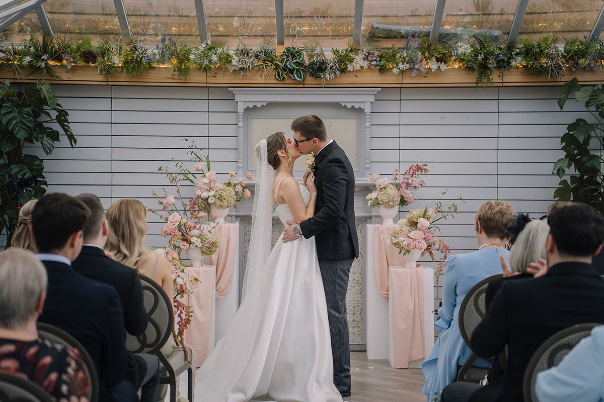 Bride and groom sharing their first kiss during the wedding ceremony in a light-filled glasshouse venue