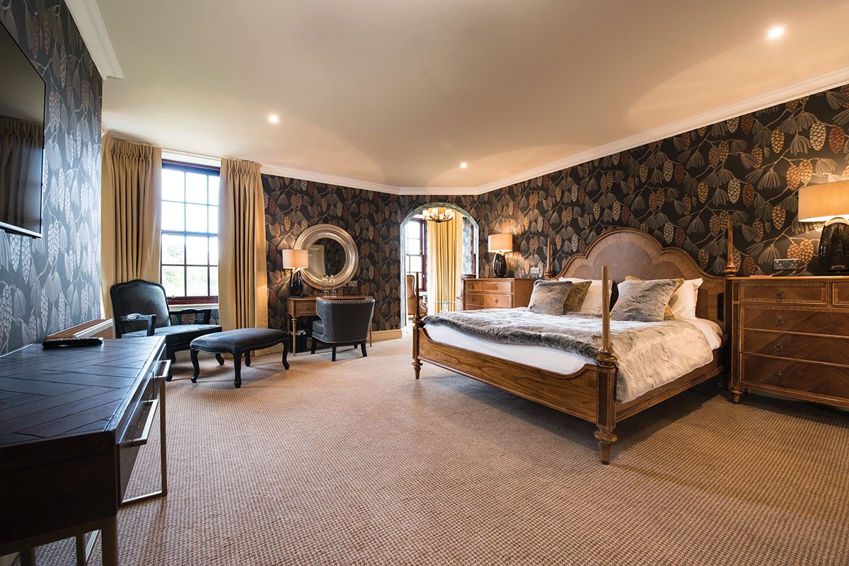 Spacious bedroom with patterned dark wallpaper, a large wooden bed, matching dressers, and armchairs by a window with gold curtains
