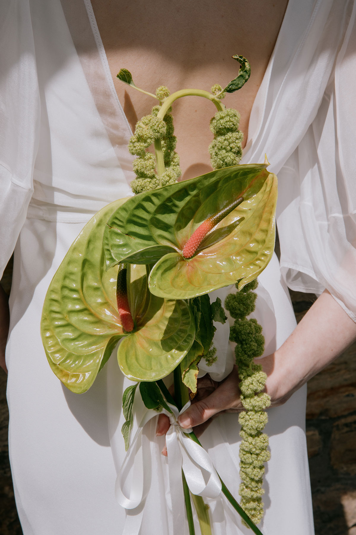 A close-up of green anthurium flowers and cascading greenery held behind a bride’s white gown, showing the open back of the dress.