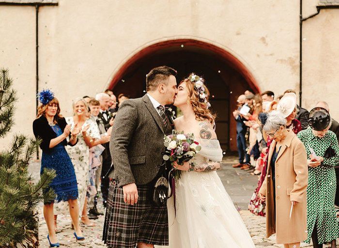 A bride holding a bouquet and wearing a flower crown and a groom in a kilt kiss in front of their venue and guests