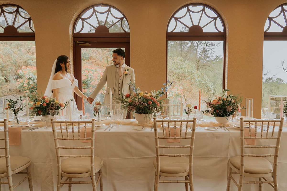 Bride and groom smiling at each other while standing beside a long wedding table decorated with pastel flowers and candles