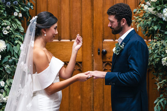 a bride holding up a wedding ring to show it to a groom while standing against a large wooden door that's flanked by a blue, white and foliage flower arrangement