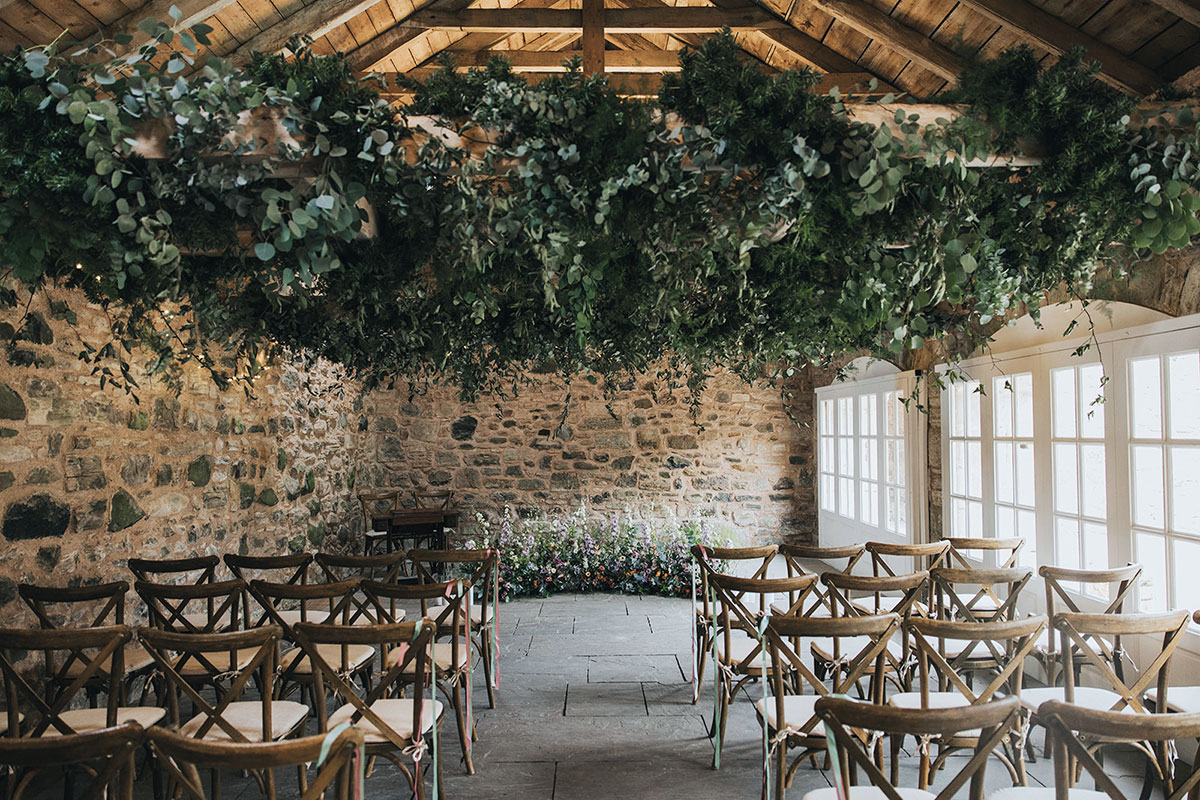 Rustic stone barn wedding ceremony set-up with hanging greenery installation, floral meadow and wooden cross-back chairs