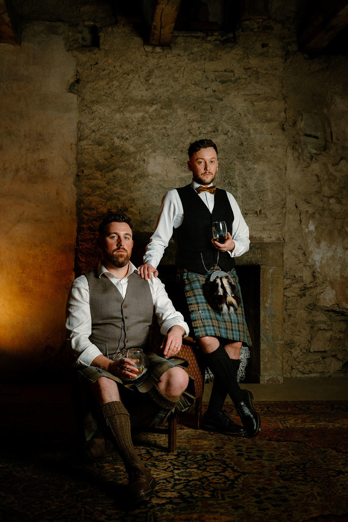 two grooms wearing kilts posing a dark textured stone room. One is seated, one is standing and they are both holding tumblers of whisky