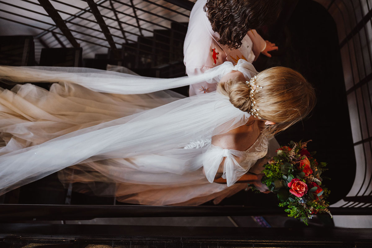 Overhead shot of two brides walking down the Engine Works staircase, the long tulle veil trailing behind and one bride carrying a colourful bouquet.