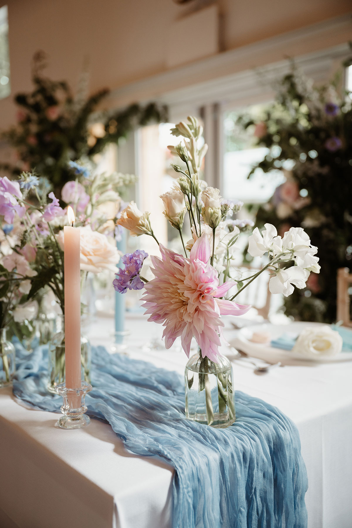 a blue crinkled fabric table runner with pastel flowers in small glass jars and pink candle in a glass holder. Table decor at Netherdale House wedding.