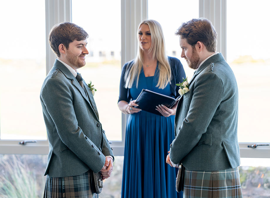 two grooms wearing kilts face each other and smile as a celebrant behind them reads from a book