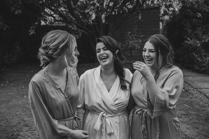 Black And White Image Of A Bride Laughing With Two Bridesmaids All Wearing Dressing Gowns In A Garden