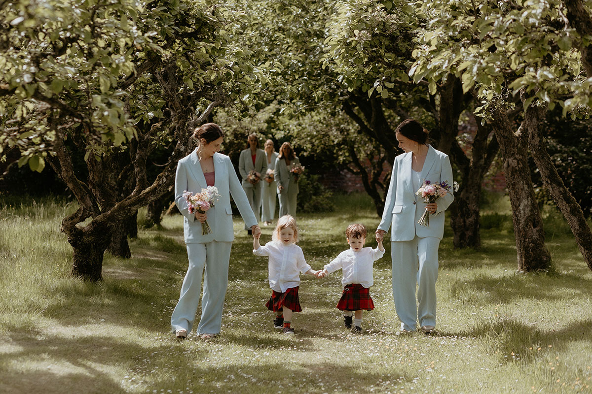 two bridesmaids wearing blue Whistles trouser suits walking down a natural outdoor aisle of trees with two toddlers wearing kilts during walled garden wedding ceremony at the Byre at Inchrya