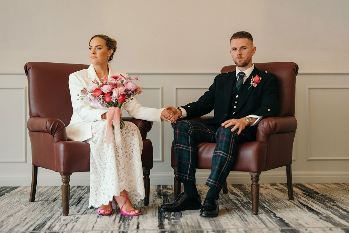 Bride in tailored white suit and groom in tartan kilt seated holding hands during wedding portrait