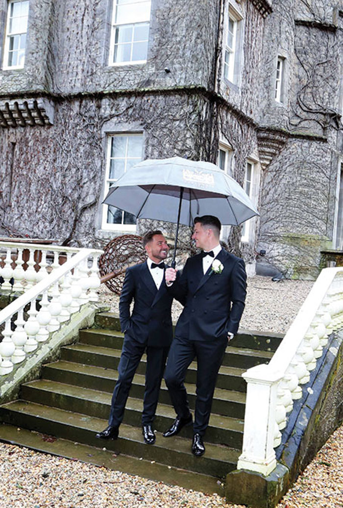 two grooms walk down a set of stone steps outside Carlowrie Castle. One holds an umbrella