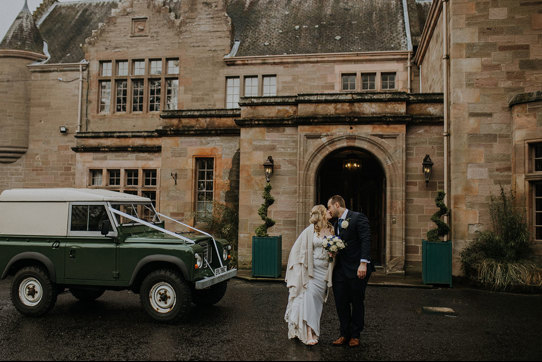 A bride and groom stand in front of a country house entrance and kiss with a green jeep next to them