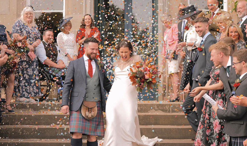 a jubilant bride and groom walking down a set of steps being showered in multicoloured paper confetti by happy guests