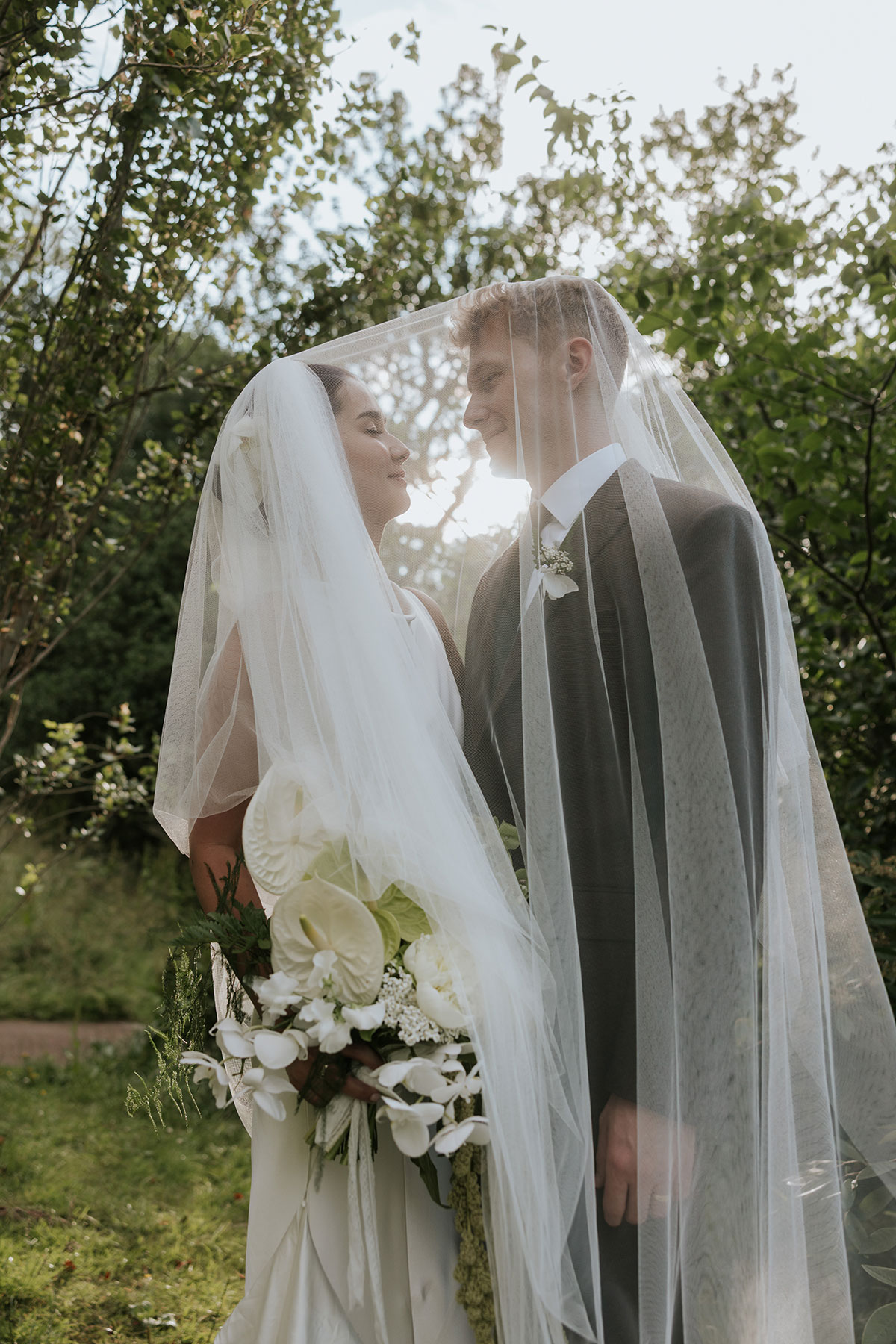 Bride and groom standing under veil in garden, sunlight shining behind them.