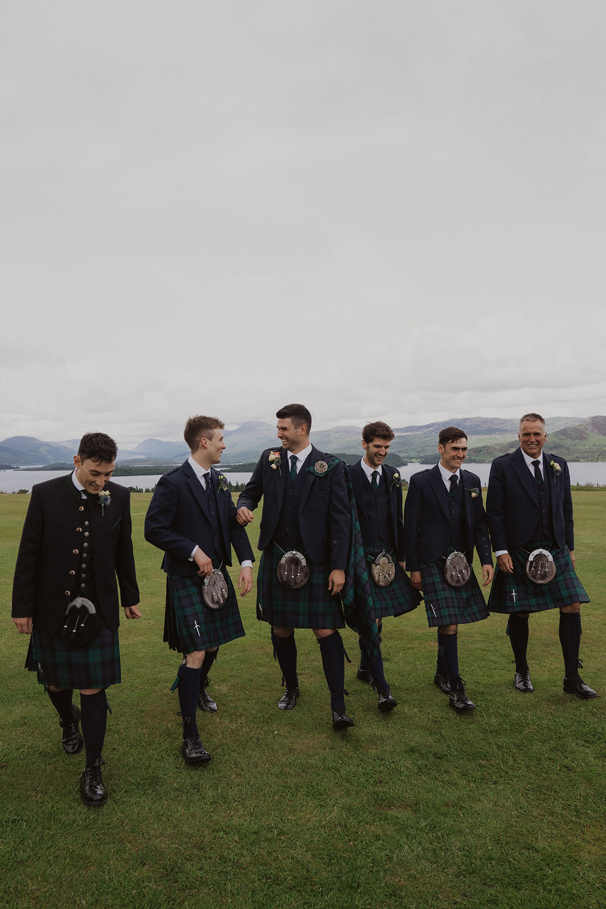 Groom and groomsmen in matching tartan kilts walking across lawn with loch and Highland hills backdrop