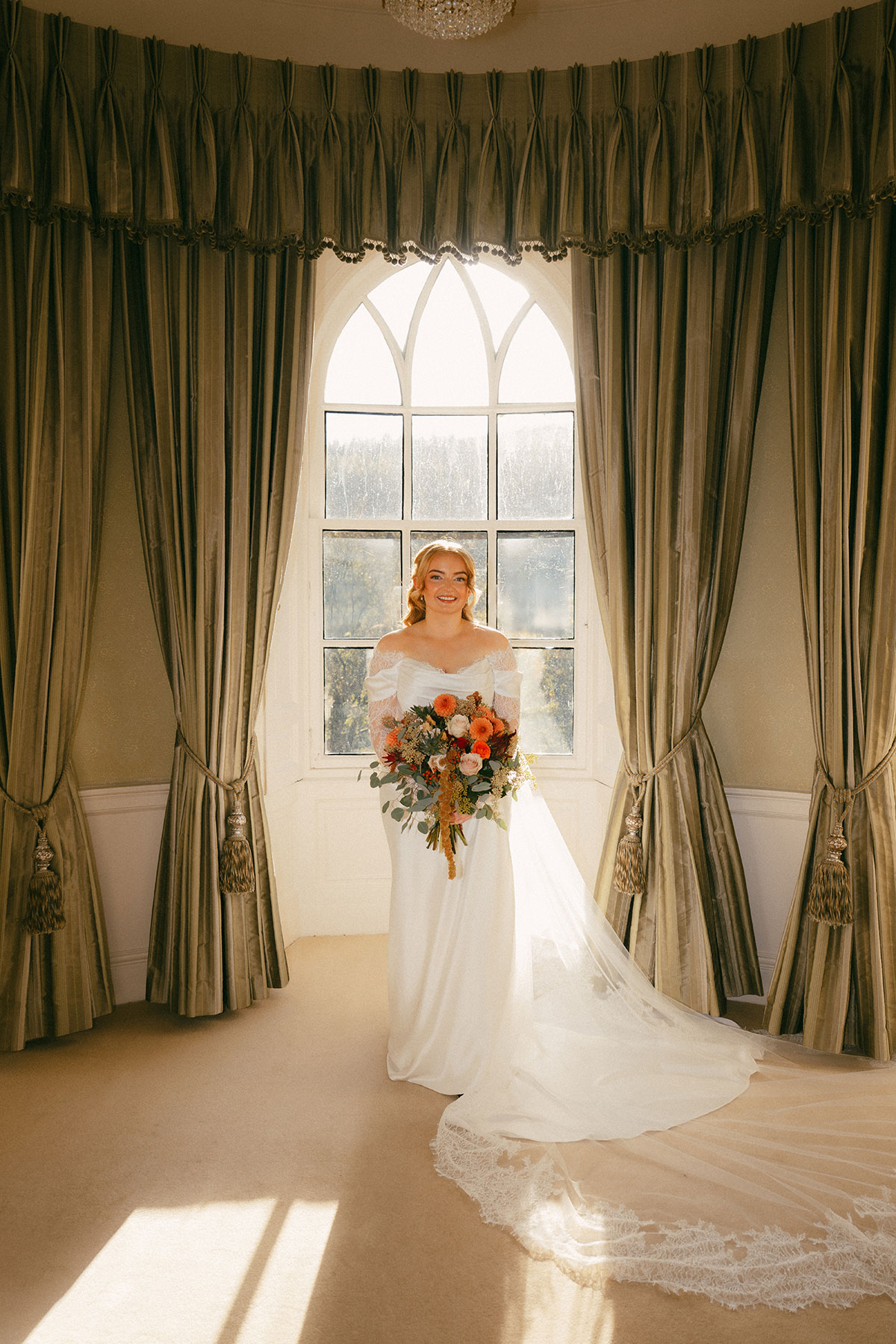 Bride in fitted off-the-shoulder wedding dress holding bouquet in front of large window at Scottish venue