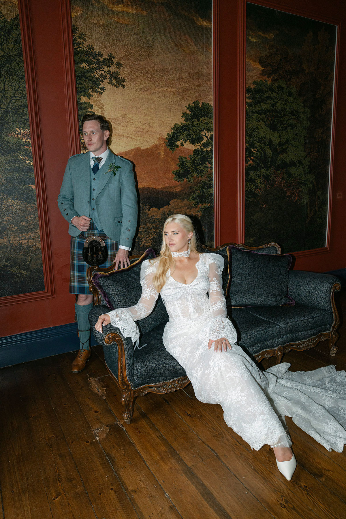 Bride in a fitted long-sleeved lace wedding dress seated on a vintage sofa beside the groom in a tartan kilt and blue jacket.