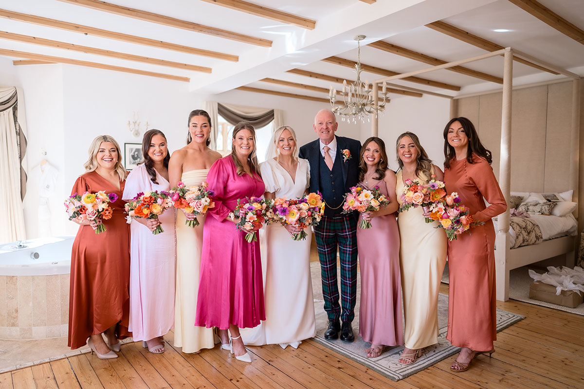 bride smiles for a photograph next to father of the bride and bridesmaids all wearing different dresses in shades of pink